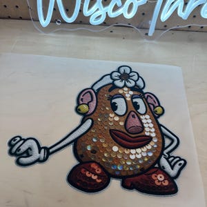 Puede incluir: Parche bordado con un personaje de dibujos animados con cuerpo de lentejuelas. El personaje tiene una flor en la cabeza, orejas rosas y zapatos rojos. El parche está sobre un soporte de color claro.