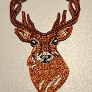 May include: An embroidered patch featuring the head of a deer with large antlers. The deer is rendered in shades of copper and brown thread with white highlights around the eyes, nose, and lower neck area, and black thread detailing for the eyes and nose.