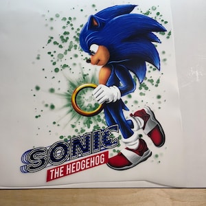Op de afbeelding: Een levendige illustratie van Sonic the Hedgehog die een gouden ring vasthoudt. Het personage is afgebeeld in zijn kenmerkende blauw met witte handschoenen en rood-witte schoenen. De afbeelding bevat de tekst "SONIC THE HEDGEHOG" in een gestileerd lettertype met een rode banner.