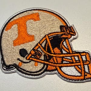 May include: An embroidered patch shaped like a football helmet in white and orange. The helmet features a large orange letter T on the side and an orange face mask. The entire design is outlined with a white border and has detailed stitching.