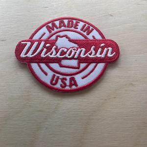 May include: Embroidered patch with a white background and a red border. The patch features the text "MADE IN Wisconsin USA" in red, with a red outline of the state of Wisconsin in the centre.