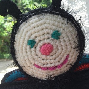 May include: A handmade crochet bee with a cream-colored face, black antennae, and a black body. The bee has a pink nose, a pink smile, and green eyes. The word "Pattern" is visible at the bottom of the image.