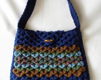 May include: A blue crocheted handbag with a brown, green and blue patterned stripe. The bag has a single strap and a wooden button closure.