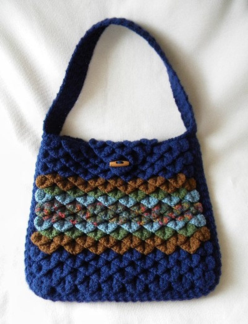 May include: A blue crocheted handbag with a brown, green, and blue patterned stripe. The bag has a single strap and a wooden button closure.