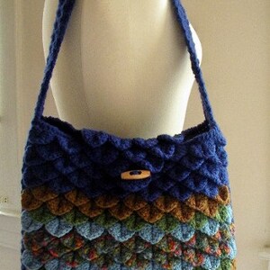 May include: A blue, green, and brown crocheted shoulder bag with a wooden button closure. The bag is made with a textured, scale-like stitch.