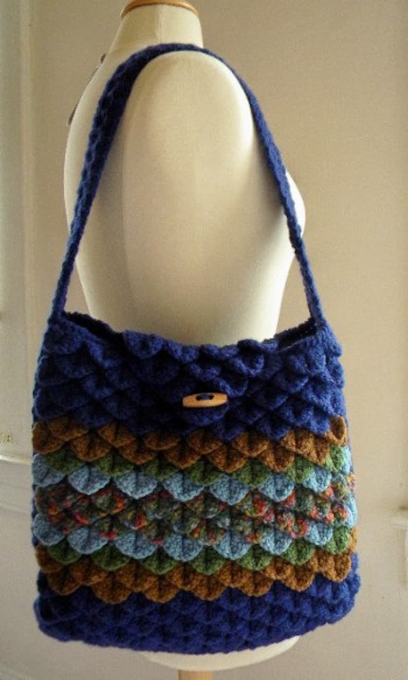 May include: A blue, green, and brown crocheted shoulder bag with a wooden button closure. The bag is made with a textured, scale-like stitch.