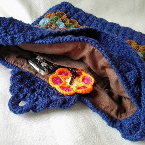 May include: A blue crocheted bag with a brown lining and a colorful crocheted flower design. The bag is open and shows a brown lining and a few items inside.