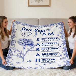 Op de afbeelding: Een witte deken met een blauw bloemenpatroon en de tekst "God zegt IK BEN". De deken heeft een grote bloem en vlinders. De tekst bevat bijbelverzen. Twee vrouwen houden de deken vast.