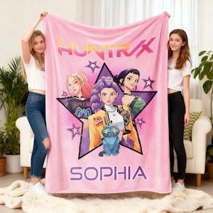 May include: Pink fleece blanket with a graphic of three stylized female figures inside a starburst. The blanket has the text "HUNTRAX" and "SOPHIA" in purple. The design includes a blue cat illustration.