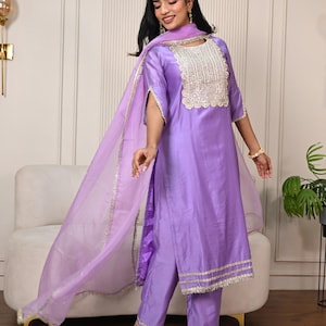 Puede incluir: Un conjunto salwar kameez de color lila. El conjunto incluye una túnica con un escote detallado, pantalones a juego y un dupatta transparente. Los bordes del atuendo están adornados con ribetes dorados. El atuendo se usa con sandalias transparentes.