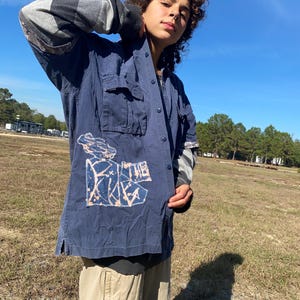 May include: A navy blue button-down shirt with two chest pockets and a graphic design on the front. The shirt has long sleeves with a grey and black patterned design on the cuffs. Khaki trousers are also visible.