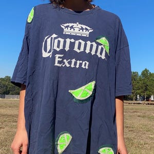 Peut inclure: T-shirt bleu marine avec le logo Corona Extra en blanc et des appliqués de tranches de citron vert. Le t-shirt a des manches courtes et une coupe décontractée. Les tranches de citron vert sont vertes avec des coutures blanches.