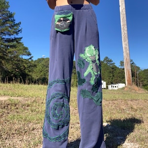 Può includere: Pantaloni blu navy con disegni applicati verdi e neri. I pantaloni presentano una tasca verde e un grande personaggio dei cartoni animati verde sulla gamba. Un disegno a spirale verde è sull'altra gamba. I pantaloni sono a gamba larga e sembrano essere fatti di un materiale morbido.