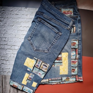 May include: Blue denim jeans with decorative patches featuring travel-themed graphics and text. The patches include images of vintage vehicles and phrases like "Hit the Road" and "Let's Get Lost". The jeans have a classic five-pocket design.