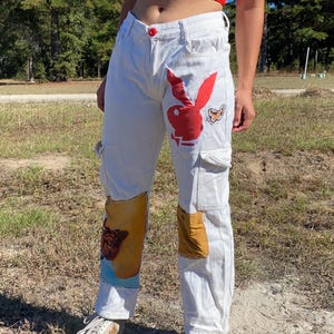 Peut inclure: Pantalon cargo en denim blanc avec logo Playboy rouge et écussons papillon. Le pantalon a plusieurs poches et une fermeture à bouton rouge. Les écussons sont dans les tons de rouge, jaune et marron.
