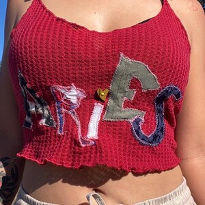 Könnte beinhalten: Ein rotes Crop-Top aus Waffelstrick mit dem Wort "ARIES" auf der Vorderseite in verschiedenen Stoffmustern appliziert. Das Top hat einen gerüschten Saum und eine kleine goldfarbene herzförmige Verzierung.