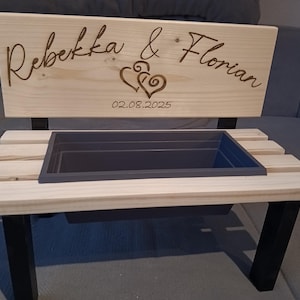 Könnte beinhalten: Eine hölzerne Pflanzbank mit einem rechteckigen grauen Behälter. Die Bank hat eine Rückwand mit den Namen "Rebekka & Florian", zwei Herzen und dem Datum "02.08.2025" eingraviert. Die Bank hat schwarze Beine und eine helle Holzoberfläche.