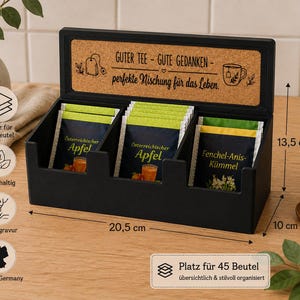 Könnte beinhalten: Eine schwarze Teebox mit drei Fächern für Teebeutel, ausgestattet mit einem Kork-Deckeleinsatz mit dem Text GUTER TEE - GUTE GEDANKEN - perfekte Mischung für das Leben. Die Box misst 20,5 cm in der Breite, 13,5 cm in der Höhe und 10 cm in der Tiefe.
