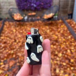 May include: Black lighter decorated with white ghost designs. The lighter has a red button and a silver metal top. The background is blurred, with fall leaves and purple flowers visible.