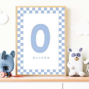 以下が含まれることがあります： 白い背景に水色の文字「O」が描かれたフレームプリント。下には「OLIVER」という名前があります。フレームは青と白のチェック柄のボーダーです。プリントは、装飾的なおもちゃが置かれた木製の棚に飾られています。