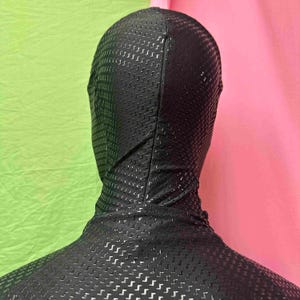 May include: A black full-body suit with a hood, featuring a textured, perforated pattern. The suit covers the head and neck, with a close fit. The background is a mix of green and pink.