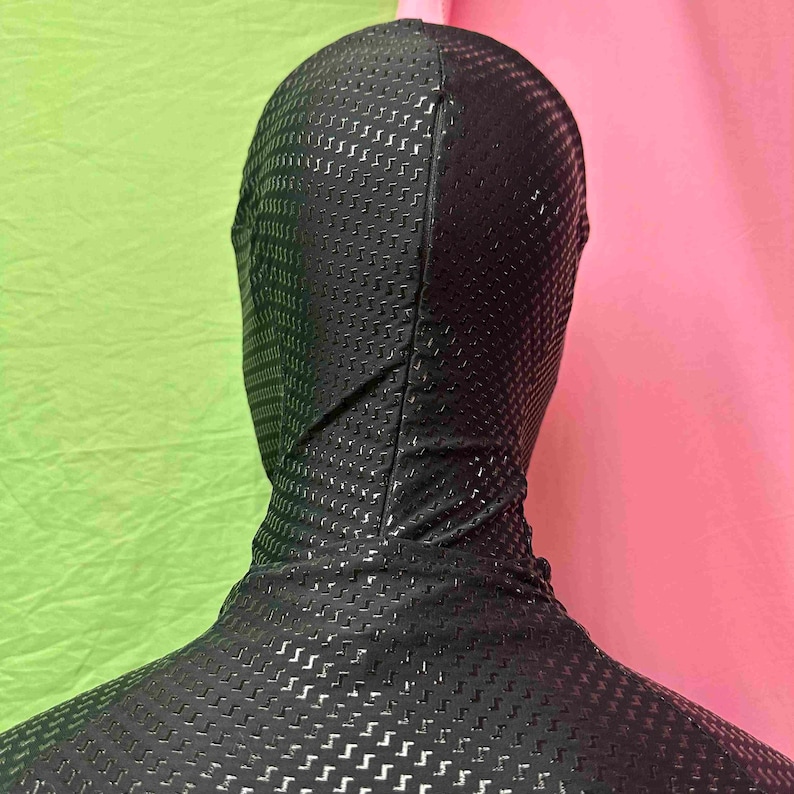 May include: A black full-body suit with a hood, featuring a textured, perforated pattern. The suit covers the head and neck, with a close fit. The background is a mix of green and pink.