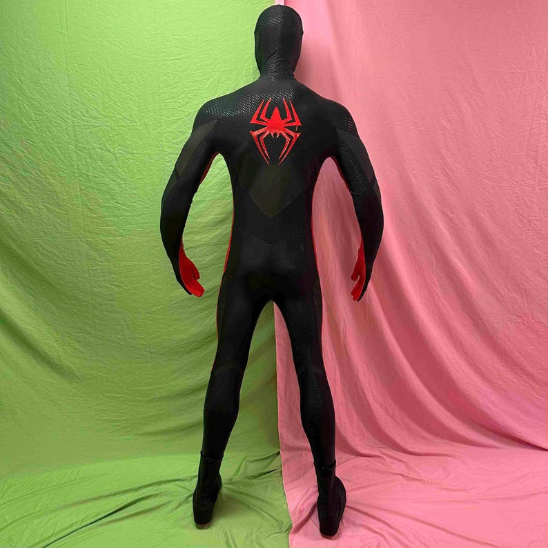 May include: Black and red superhero costume featuring a spider emblem on the back. The full-body suit has a textured pattern and includes a hood, gloves, and boots. The costume is displayed against a pink and green backdrop.