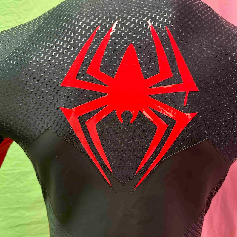 May include: Black and red superhero costume with a large red spider emblem on the back. The costume has a textured, carbon fibre-like pattern on the upper back and shoulders. The design includes a sleek, form-fitting silhouette.