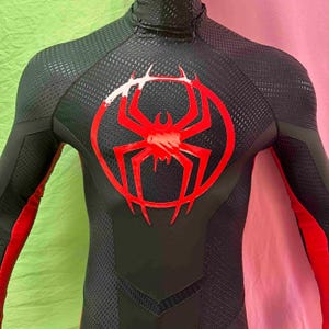 May include: Black and red superhero costume featuring a red spider emblem on the chest. The suit has a textured pattern and red accents on the arms. The costume has a high collar.