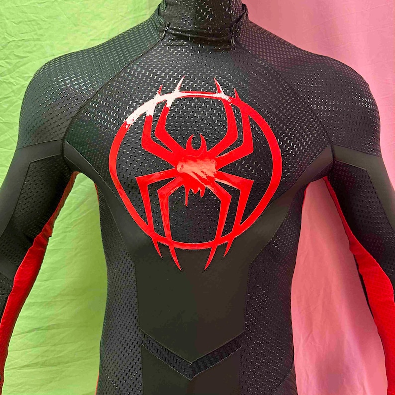 May include: Black and red superhero costume featuring a red spider emblem on the chest. The suit has a textured pattern and red accents on the arms. The costume has a high collar.