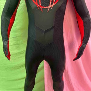 May include: A black and red bodysuit with a textured pattern. The suit has red accents on the arms and chest, with a detailed design on the upper back. The gloves are black with red detailing. The suit appears to be made of a flexible material.