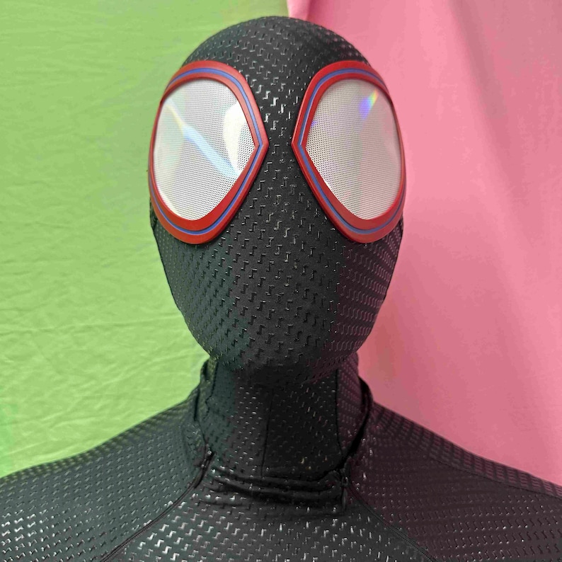 May include: A black superhero mask with red and blue trim around the eye openings. The mask has a textured pattern. The eye lenses are white. The background is green and pink.