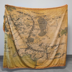 May include: A large, rectangular tapestry featuring a detailed map of Middle-earth. The map is rendered in shades of brown and tan, with black text and illustrations. The tapestry has a vintage, aged appearance, with the names of locations like Rohan and Mordor.