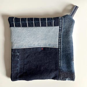 May include: A patchwork denim pouch with a striped top flap and a blue denim strap. The pouch is made of different shades of blue denim and has a unique, handmade look.