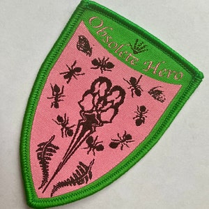 May include: A pink and green embroidered patch with a skeletal hand and ants. The text "Obsolete Hero" is written in a green banner at the top of the patch.