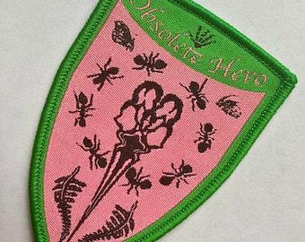May include: A pink and green embroidered patch with a skeletal hand and ants. The text "Obsolete Hero" is written in a green banner at the top of the patch.