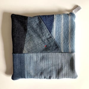May include: Square denim potholder with a patchwork design. The potholder features various shades of blue denim, including a dark blue square, a triangle, and a striped section. A small red stitch is visible. A gray loop is attached for hanging.