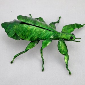 Puede incluir: Una escultura de insecto palo de metal verde con patas y alas detalladas. El insecto est&aacute; en posici&oacute;n de caminar con las patas extendidas.