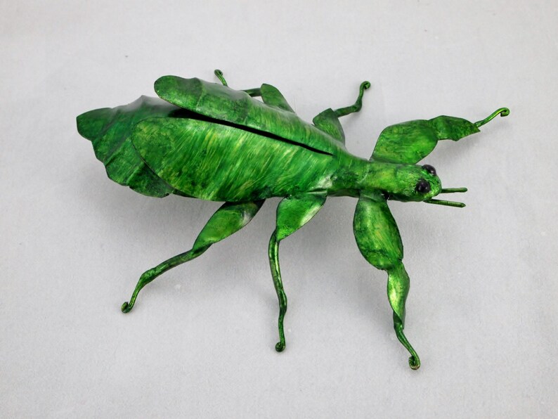 Puede incluir: Una escultura de insecto palo de metal verde con patas y alas detalladas. El insecto est&aacute; en posici&oacute;n de caminar con las patas extendidas.