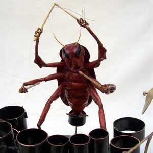 May include: A red and black bug sculpture playing a drum set. The bug is holding drumsticks in its front legs and has a red body with black eyes. The drum set is made of metal and has a variety of drums and cymbals.