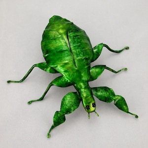 Puede incluir: Una escultura de metal verde de un insecto palo con alas texturizadas detalladas y ojos negros.