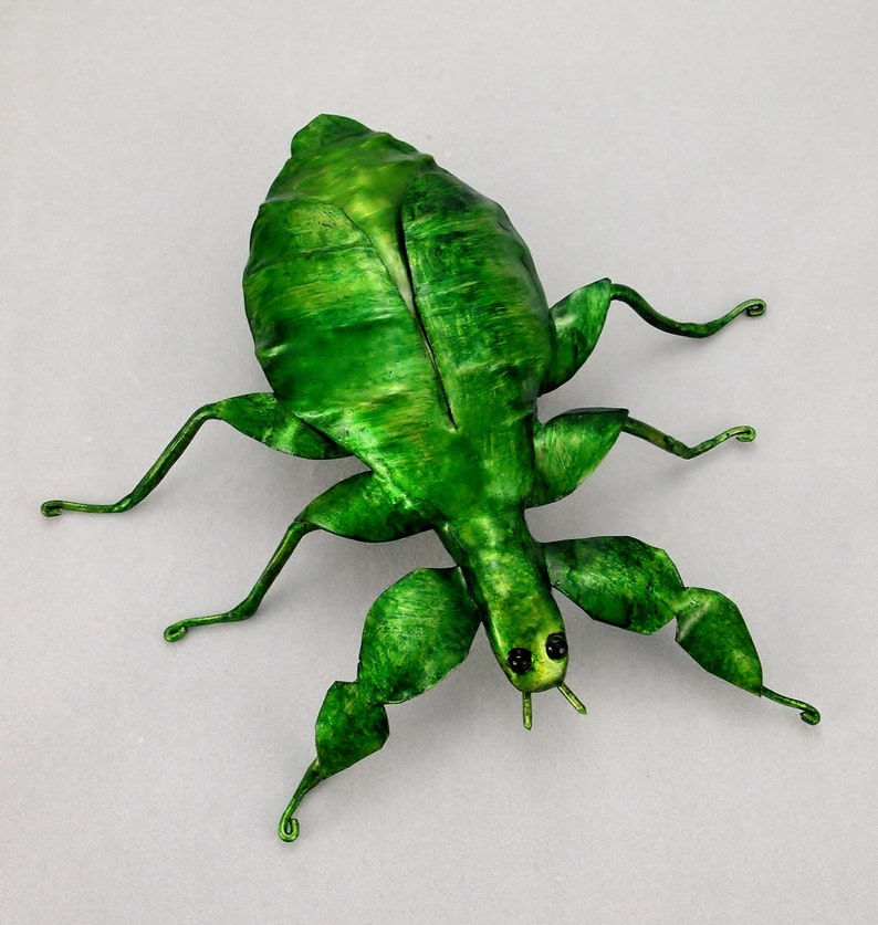 Puede incluir: Una escultura de metal verde de un insecto palo con alas texturizadas detalladas y ojos negros.