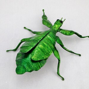 Puede incluir: Una escultura de metal verde de un insecto palo con patas largas y un cuerpo con forma de hoja.