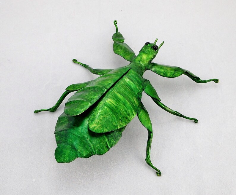 Puede incluir: Una escultura de metal verde de un insecto palo con patas largas y un cuerpo con forma de hoja.