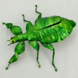 Puede incluir: Una escultura de metal verde de un insecto palo con patas largas y un cuerpo detallado. La escultura est&aacute; hecha para parecer un insecto real.