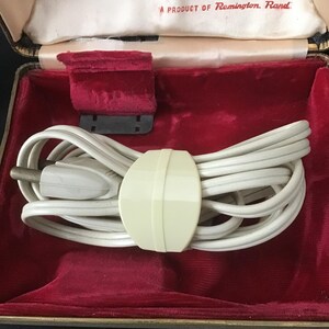 May include: A vintage Remington Rand electric shaver in a black and gold case with a red velvet interior. The case contains a white cord, a cream-coloured cord holder, and a red velvet pad. The text "A PRODUCT OF Remington Rand" is visible.