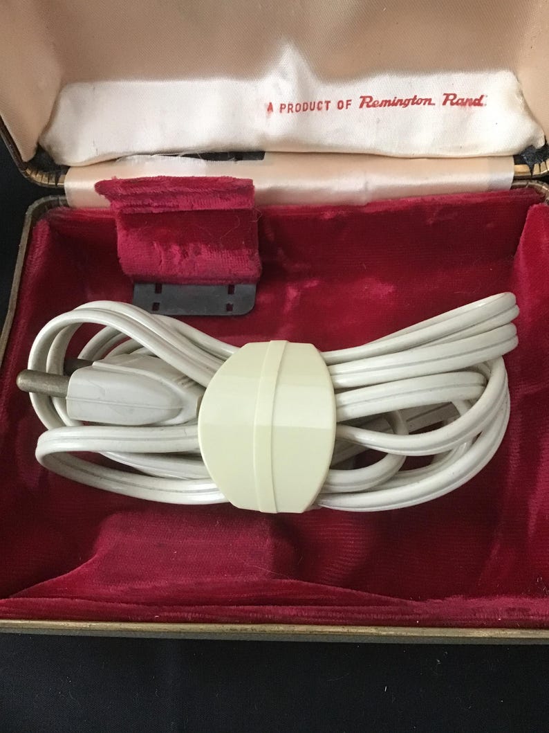 May include: A vintage Remington Rand electric shaver in a black and gold case with a red velvet interior. The case contains a white cord, a cream-coloured cord holder, and a red velvet pad. The text "A PRODUCT OF Remington Rand" is visible.