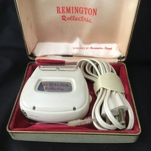 May include: A vintage Remington Rollectric electric shaver in its original case. The white shaver has a silver blade and is housed in a red velvet-lined case with the Remington Rollectric logo in red. A cleaning brush and power cord are included.