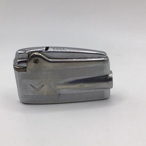 Peut inclure: Un briquet vintage en métal argenté avec un corps rectangulaire et un dessus articulé. Le briquet a une surface texturée avec un motif en chevrons sur le devant. Un objet de collection classique.