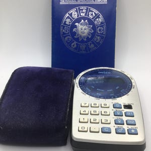 Könnte beinhalten: Ein Vintage Astro persönlicher astrologischer Computer von Kosmos. Das Gerät hat ein silbernes und schwarzes Gehäuse mit blauen Akzenten und einer Tastatur. Ein blaues Büchlein und ein dunkelblaues Samtetui sind ebenfalls vorhanden.